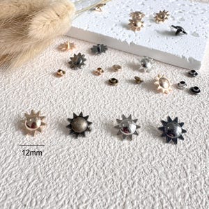 May include: An assortment of sun-shaped metal rivets in various finishes, including gold, silver, and antique bronze. The rivets are approximately 0.5 inches (12mm) in diameter and are displayed on a textured white surface. Additional small rivets are also present.