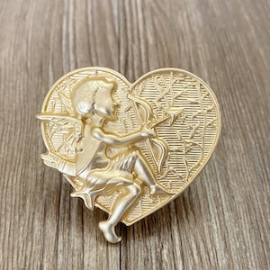 May include: A gold-coloured heart-shaped brooch featuring a detailed design of a cupid holding an arrow. The cupid is sitting on a heart with a vine pattern.