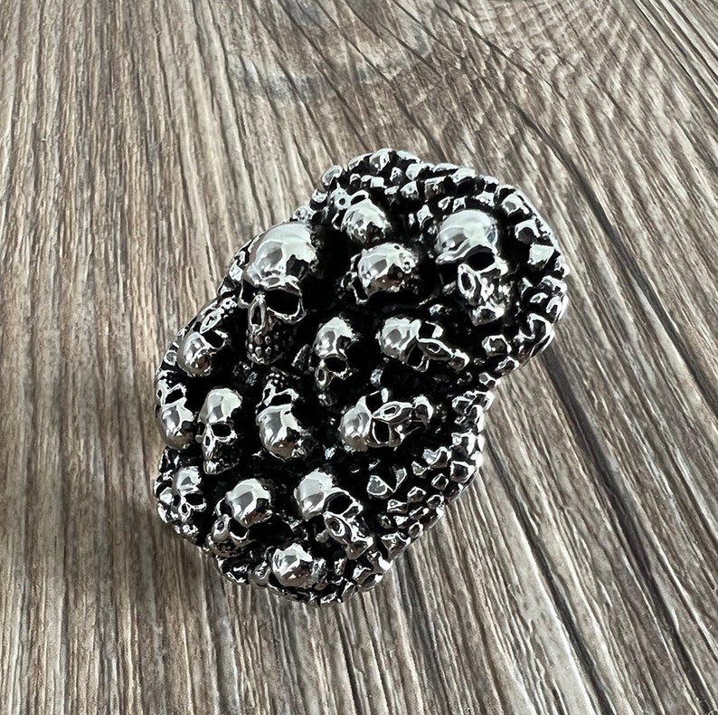 K&ouml;nnte beinhalten: Eine silberfarbene, rechteckige G&uuml;rtelschnalle, dicht bedeckt mit kleinen Sch&auml;deln. Die Sch&auml;del weisen detaillierte Merkmale auf und sind in einem geclusterten Muster angeordnet. Die Schnalle hat eine polierte, reflektierende Oberfl&auml;che und ein gotisches Design.