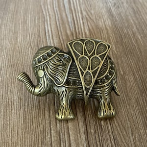 以下が含まれることがあります： 複雑なディテールが施されたブロンズ色の象の置物。象は鼻を上げており、背中に装飾的なデザインが施されています。