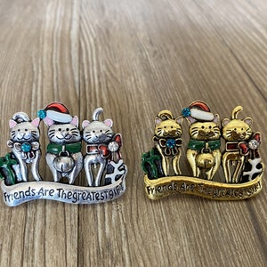 Puede incluir: Tres figuritas de gatos de plata y oro que llevan gorros de Papá Noel y sostienen regalos. Los gatos están de pie sobre una pancarta que dice "Friends Are The Greatest Gift".