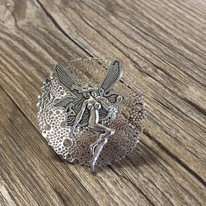 May include: A silver fairy figurine with wings, standing on a circular, silver, openwork base. The base has a delicate, lacy design.