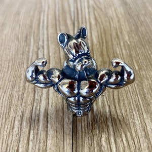 Op de afbeelding: Een zilverkleurige metalen deurklink in de vorm van een gespierd figuur met een cartoonachtig gezicht. De figuur buigt zijn biceps.