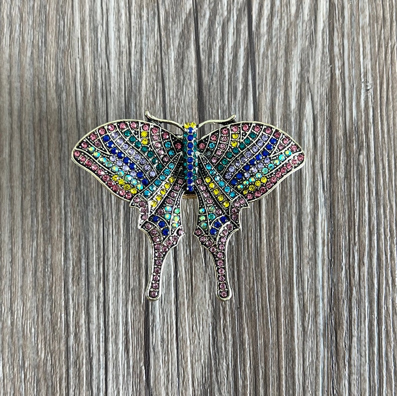 May include: A colourful butterfly brooch with a gold finish. The butterfly is covered in rhinestones of various colours, including pink, blue, green, and yellow.