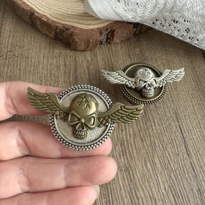 Puede incluir: Dos adornos metálicos en forma de calavera con alas. Uno tiene una combinación de colores plateado y dorado, el otro una combinación de colores bronce y plateado. Los adornos son redondos con un borde de cuentas y están sobre una superficie de madera.