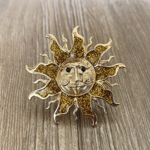 Può includere: Una spilla a forma di sole dorata con finitura glitter e un viso con occhi neri.