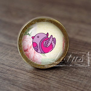 May include: A round, brass-colored cabinet knob with a glass insert featuring a pink and purple bird illustration on a floral background.