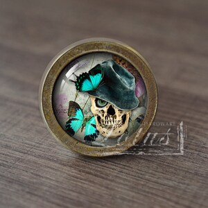 Puede incluir: Un pomo de armario redondo de color latón con un inserto de vidrio que presenta una calavera con un sombrero azul y rodeado de mariposas turquesa.
