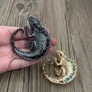 Może przedstawiać: Dwa broszki w kształcie jaszczurki, ozdobione kamieniami szlachetnymi, jedna w kolorze czarnym z czarnymi i zielonymi kamieniami, a druga w kolorze złotym z zielonymi kamieniami.