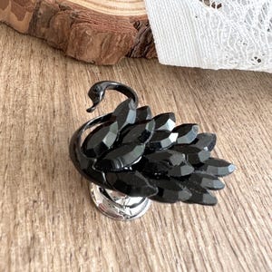 Peut inclure: Broche en forme de cygne noir, avec une base argentée. Le corps du cygne est fait de pierres noires facettées, et son cou est courbé avec élégance. La broche est posée sur une surface en bois.
