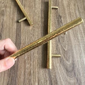 Peut inclure: Trois poignées de tiroir en métal martelé doré avec une finition brossée. Les poignées sont de différentes longueurs et ont un design simple.