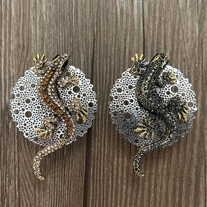 Puede incluir: Un par de pendientes con un diseño de lagarto enjoyado sobre una base circular texturizada. El lagarto del pendiente izquierdo es marrón y dorado, mientras que el lagarto del pendiente derecho es negro y dorado.