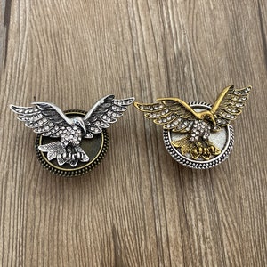 Pode incluir: Dois pingentes de pressão em forma de águia com strass. Um pingente é prateado com fundo marrom e o outro é dourado com fundo marrom.