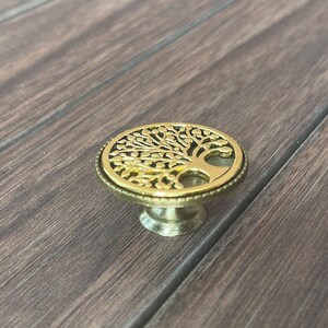 May include: A gold-toned metal cabinet knob with a tree of life design. The knob has a round base and a raised tree design with intricate details.