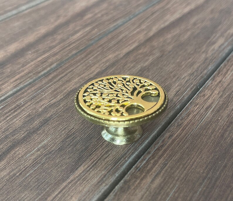 May include: A gold-toned metal cabinet knob with a tree of life design. The knob has a round base and a raised tree design with intricate details.