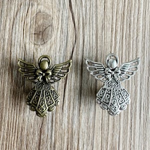 Puede incluir: Dos encantos de ángel, uno plateado y otro bronce, con detalles intrincados y un lazo en la parte superior. Los encantos están sobre un fondo de madera.