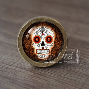 Könnte beinhalten: Ein runder, antiker Messing-Schrankknauf mit einem Glaseinsatz, der ein farbenfrohes Totenkopf-Design im Stil des Día de Muertos zeigt. Der Schädel ist weiß mit schwarzen Umrissen und roten und orangefarbenen Akzenten.