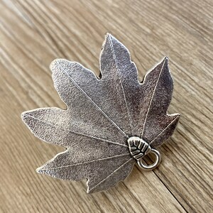 Pode incluir: Encanto de folha de metal prateado com uma superf&iacute;cie texturizada detalhada e um pequeno la&ccedil;o na parte inferior.