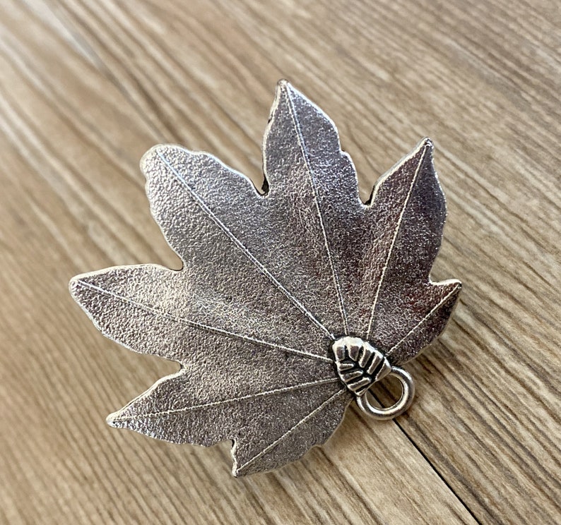 Pode incluir: Encanto de folha de metal prateado com uma superf&iacute;cie texturizada detalhada e um pequeno la&ccedil;o na parte inferior.