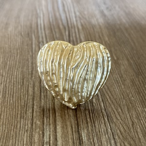 Puede incluir: Un objeto en forma de corazón de color dorado con una superficie texturizada. El corazón tiene un patrón acanalado y el diseño general es brillante. El objeto es probablemente un artículo decorativo o una joya.