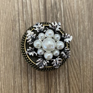 May include: A round, bronze-coloured button with a silver snowflake design. The snowflake is adorned with white pearl-like beads.