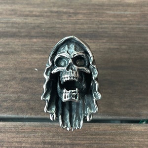 May include: A silver ring with a detailed skull design, featuring an open mouth and a hooded cloak. The skull has visible teeth and eye sockets, with the hood draping around the head. The ring is set against a wooden background.