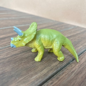 May include: A green and yellow Triceratops dinosaur toy with blue horns and a blue beak. The toy is made of plastic and is on a wooden surface.