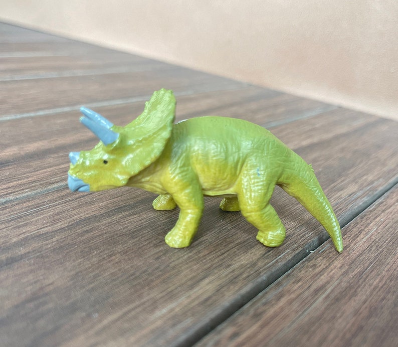 May include: A green and yellow Triceratops dinosaur toy with blue horns and a blue beak. The toy is made of plastic and is on a wooden surface.