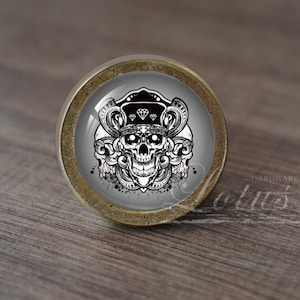 Könnte beinhalten: Ein runder Schrankknopf mit Totenkopf-Design. Der Knopf hat eine klare, gewölbte Glasfläche mit einem schwarz-weißen Totenkopf, der eine Krone trägt. Der Metallsockel hat eine Bronzefinish. Dekorative Hardware.