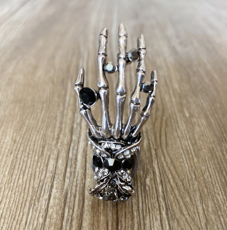 K&ouml;nnte beinhalten: Ein silberner Skeletthandring mit schwarzen und klaren Strasssteinen. Der Ring zeigt eine detaillierte Skeletthand mit einem Sch&auml;del an der Basis.