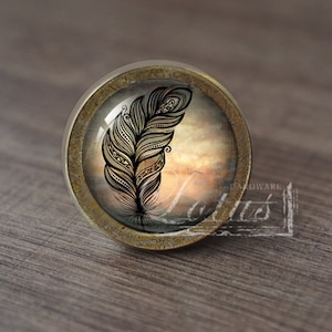 May include: A round, brass-colored cabinet knob with a glass insert featuring a black and white patterned feather design on a cloudy, orange and brown background.