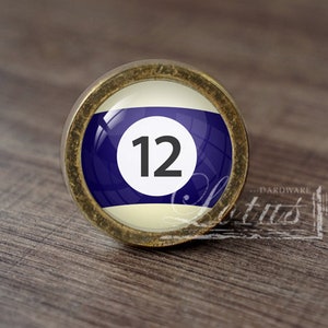 Puede incluir: Un pomo de latón con un inserto de vidrio que presenta una bola de billar blanca con el número 12 en negro.
