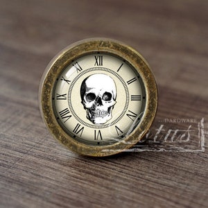 May include: A round, brass-colored cabinet knob with a white face and black Roman numeral clock face. The clock face features a black and white skull illustration in the center.