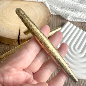 Könnte beinhalten: Nahaufnahme eines Satzes goldfarbener Schrankgriffe mit gehammerter Textur. Die Griffe haben eine zylindrische Form mit abgerundetem Ende und kleinem, rechteckigem Sockel. Der Hintergrund umfasst eine Holzscheibe und ein weißes Dekorationselement.
