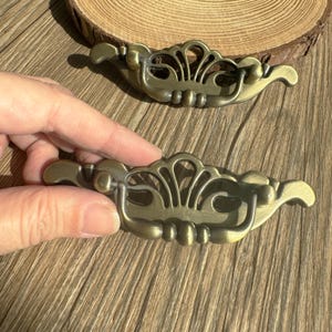 May include: Two antique brass drawer pulls with an ornate, symmetrical design. Each pull features a curved handle and decorative filigree details, perfect for adding a vintage touch to furniture. The pulls are shown on a wooden surface.