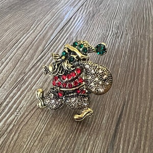 Könnte beinhalten: Eine goldfarbene Weihnachtsmann-Brosche mit roten und klaren Strasssteinen. Der Weihnachtsmann trägt einen grünen Hut mit einem grünen Stein oben drauf. Er trägt einen Sack mit Geschenken.