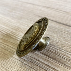 May include: A round, brass cabinet knob featuring an eagle design. The knob has a decorative border and a textured surface.