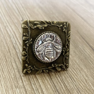 May include: A square, ornate, metal, decorative piece with a floral design. The center features a silver bee with a detailed design.