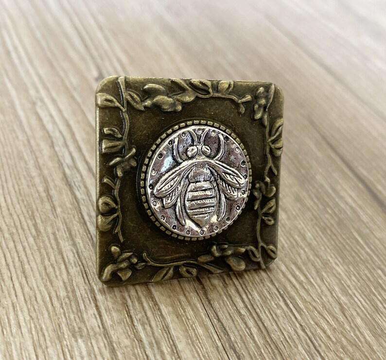 May include: A square, ornate, metal, decorative piece with a floral design. The center features a silver bee with a detailed design.