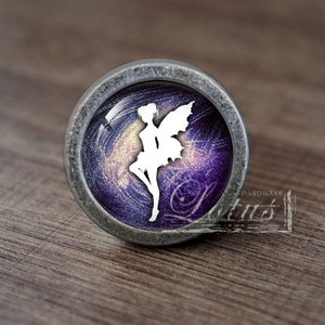 May include: A circular cabinet knob with a silver-coloured metal surround. The glass insert displays a white fairy figure on a swirling purple and gold backdrop. The knob has a vintage appearance.