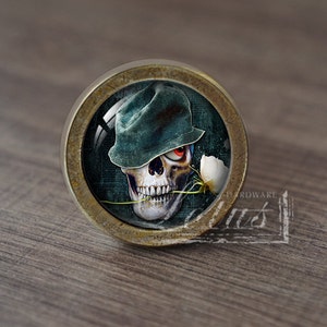 Pode incluir: Um botão de armário redondo de cor latão com um inserto de vidro que apresenta uma caveira com um chapéu verde e segurando uma flor branca. A caveira tem um olho vermelho e um sorriso de dentes.