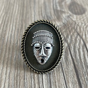 Op de afbeelding: Een zilveren stammesmasker met een kralenhoofdtooi wordt getoond in een rond, bronskleurig frame met een touwachtige rand.