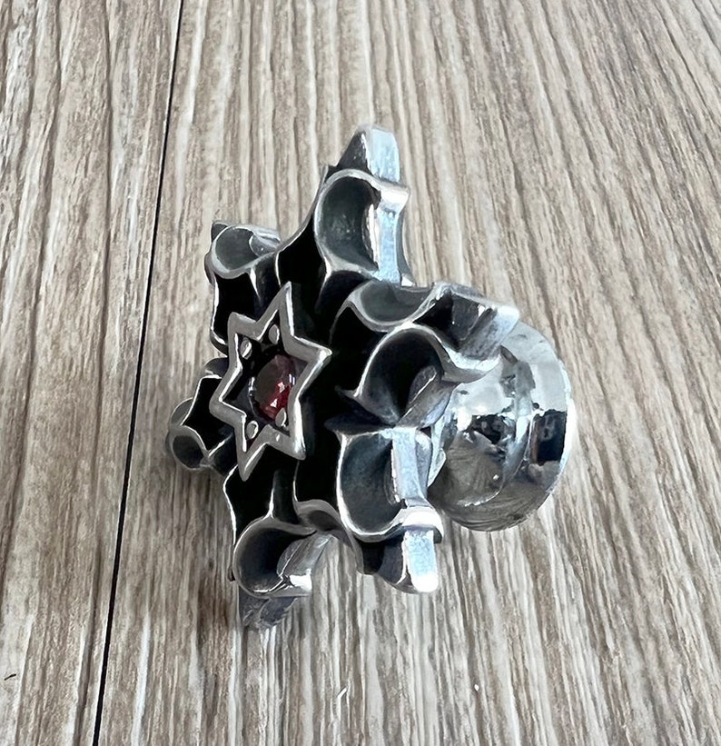 Puede incluir: Un anillo de plata con un dise&ntilde;o detallado que presenta una estrella con una piedra preciosa roja en el centro. El anillo est&aacute; colocado sobre una superficie de madera.