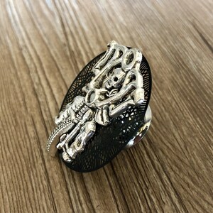 Op de afbeelding: Een zilverkleurige metalen ring met een zwarte achtergrond. De ring heeft een gedetailleerd ontwerp van een skelet dat een motorfiets rijdt. De motorfiets heeft een schedel als koplamp en de woorden "Ride or Die" zijn zichtbaar op de ring.