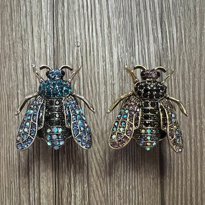 Könnte beinhalten: Zwei dekorative Broschen in Bienenform mit juwelenbesetzten Flügeln. Eine Brosche hat blaue und schillernde Flügel, die andere hat violette und schillernde Flügel.