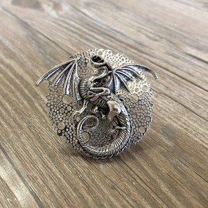 May include: Silver dragon ring with a detailed design. The dragon is curled around a circular base with a lacy pattern.