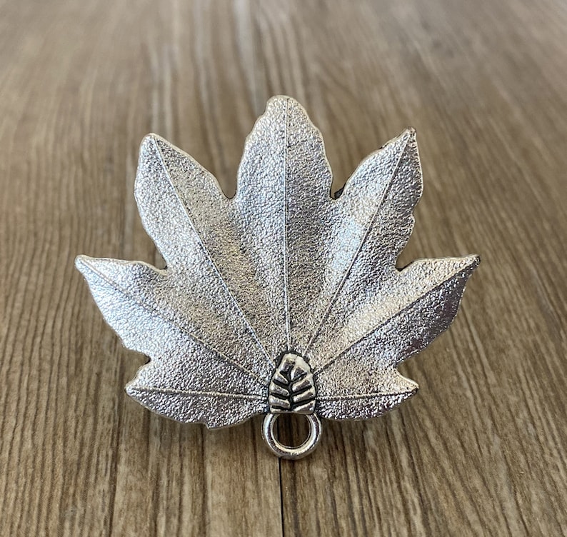 Pode incluir: Encanto de folha de metal prateado com uma superf&iacute;cie texturizada detalhada e um pequeno la&ccedil;o na parte inferior.