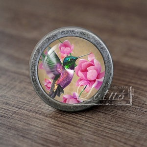 Puede incluir: Un pomo plateado con un inserto de vidrio que presenta un colibrí en vuelo con flores rosas. El colibrí es verde, morado y negro con un pico largo. Las flores son rosas y blancas.