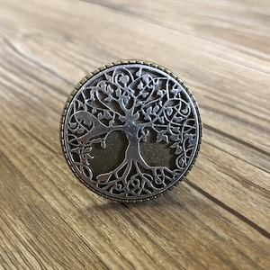 May include: A silver and bronze circular pendant featuring a detailed tree of life design. The tree has intricate branches and roots, and the background is a textured bronze color.