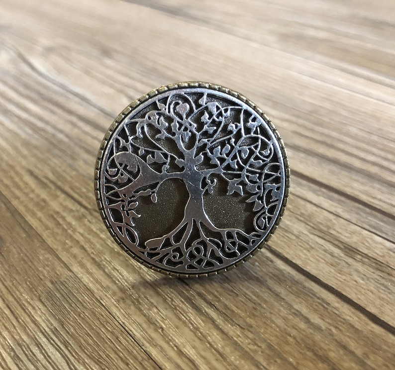 May include: A silver and bronze circular pendant featuring a detailed tree of life design. The tree has intricate branches and roots, and the background is a textured bronze color.